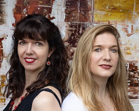 Photo of who white women, one dark haired, one blonde, standing back to back