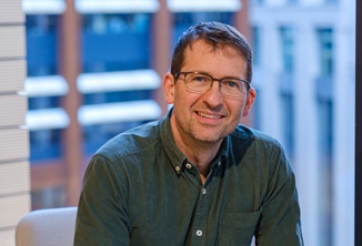 Image of a white man with glasses and a grey shirt in a blue building