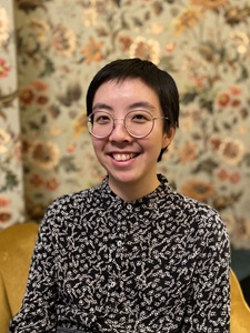 Photo from chest upwards of author in black and white top and short hair against floral wallpaper