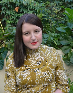 Author photo from chest upwards of a white woman with dark hair and a textured yellow top against leaves