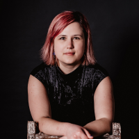Headshot of a white woman with red hair leaning on the back of a chair with a black top against black