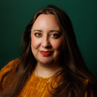 Headshot of a white woman with long dark hair and a yellow jumper against green