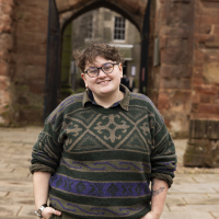 Image of a white person in a green jumper before a brown gateway