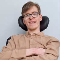 A white man wearing a brown shirt sits in a wheelchair in front of a pale blue background