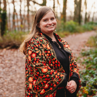 Alyssa, a white woman with blonde hair and a big smile stands in autumnal woodland in a black blouse and thick patterned wrap in bright autumnal colours.