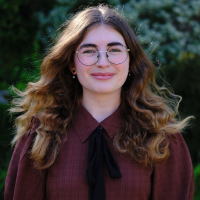 A white woman with wavy brunette hair and round glasses, stands before a hedge smiling in a maroon shirt with a long black bow tied at the collar