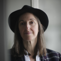 Frances stands half in shadow wearing a hat. She has shoulder length brown hair and a tartan tailored jacket,