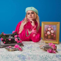 Image of white woman with blonde hair in a light blue beret wearing a hot pink blouse with poofy long sleeves and silk scarf with grey pearls sat behind a table with a damask table cloth in front of a blue background surrounded by roses