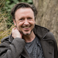 A white man stand in front of a tree and long grass in a grey shirt and coat. 