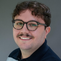 A white man, with short,dark, curly hair and a moustache smiles at the camera. He is wearing round horn rimmed glasses, and a light blue and navy striped polo shirt.