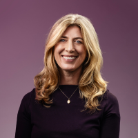 A white woman with a big smile and shoulder length wavy blonde hair sits in front of a purple background.