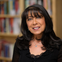 A woman with dark shoulder length hair, wearing a black top, sits smiling in front of a bookcase in the blurred background.