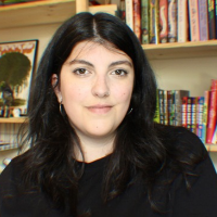 A white person with long dark hair in a black top sits in front of bookshelves smiling at the camera.
