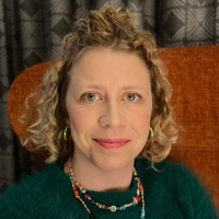 A white woman with chin length curly blonde hair, wearing a dark jumper sits in an orange chair smiling.
